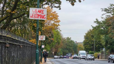 White signs with red painted messages saying 'we are welcoming' and 'we are kind' are hung on green lamp-posts lining a road in front of a black metal fence. There are cars in the distance and a man walking along the pavement. The trees are yellow and orange and green.