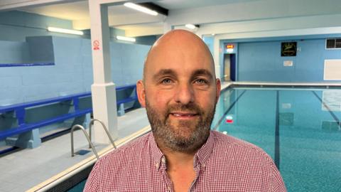 Julian Frost, 43, has a bald head and grey and black stubbly beard and moustache. He is wearing a red and white gingham. Behind him is a swimming pool.