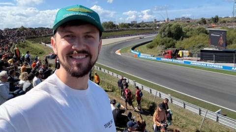 Brandon Burgess is taking a selfie. He has dark brown hair and a short beard. He is wearing a green cap which has a golden logo and the words 'Formula One Team' on the front. He also wears a white round neck short sleeved t shirt. He is standing in a crowd of people on teh side of a tarmac race track.