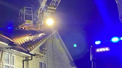 An Aerial Ladder Platform perches just above the roof of a hotel building, by which point some of its slats have been taken off so firefighters can check for hotspots. There are bright blue lights to the right of the image which are coming from the top of a fire engine. 