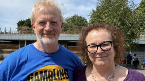 A man in a blue T-shirt and a woman in a purple top. He has short white and blonde hair and she has curly brown hair