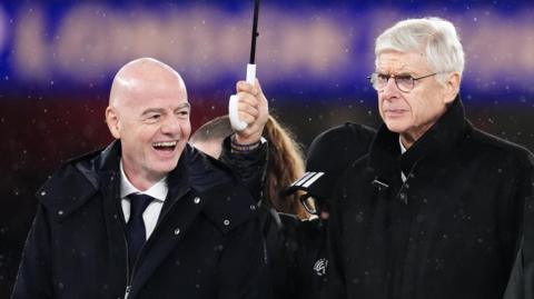 Gianni Infantino is smiling wearing a black coat and Arsene Wenger walks slightly behind wearing a black coat with a stern expression