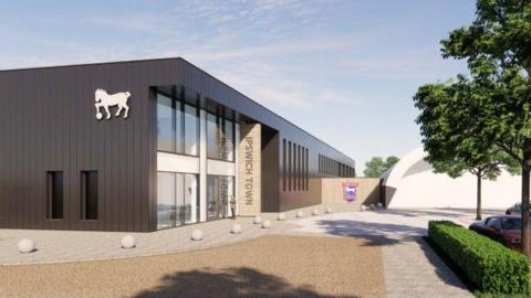 Modern brown two-storey building with glass entrance frontage. There is a logo on the side of the building featuring a Suffolk punch horse with a football. There are small stone circles on the ground. The existing indoor dome is visible to the right, behind a tree and hedge.