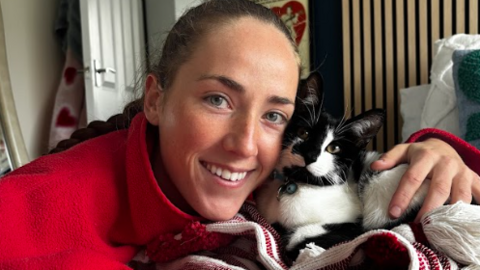 A woman with tied-back brown hair and a red top holding a black and white cat on a blanket