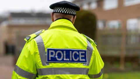 A police officer, seen from behind, walking down a residential street. He is wearing a high vis jacket and a police hat. 