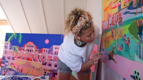 Ruth Mulvie leaning over painting one of her brightly coloured works. The picture is of Saltdean Lido, people in the water, sitting on the side of the pool, a dog in the foreground, a ball.