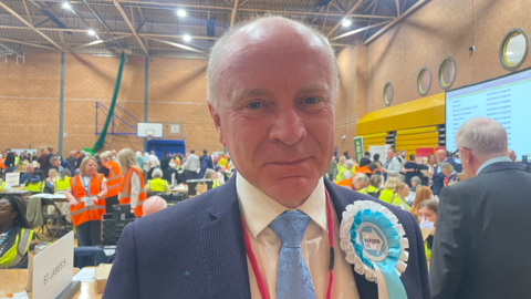 A man is wearing a blue jacket and tie, a light shirt and light blue and white rosette. He is close to the camera and many people are inside behind him in the background.