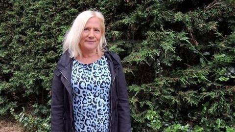 Fiona Rosevear wears an animal print black and white top and black annorak over the top. She stands in front of greenery.