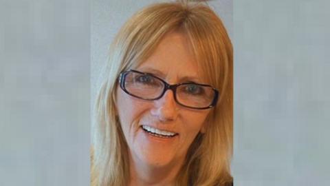 A smiling woman with blonde shoulder-length hair wearing dark-framed glasses