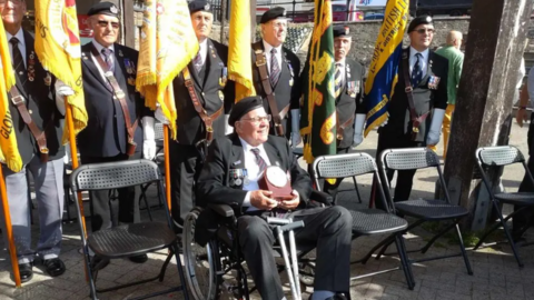 Roy Mills in a wheelchair dressed in a suit and an army cap with his medals on the lapel. Behind him six men are in similar dress, holding large yellow regimental flags.