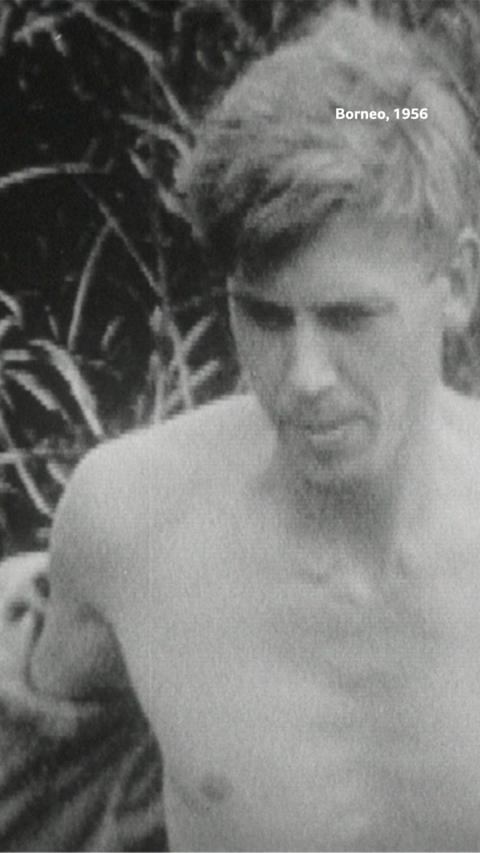 A close-up of Sir David Attenborough topless walking through a forest