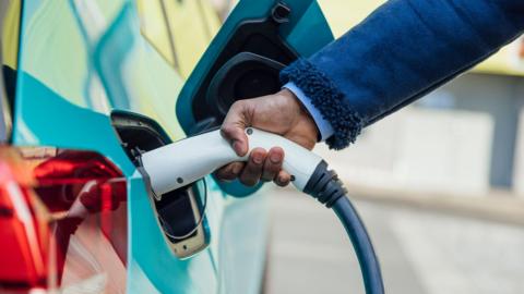 A side view close up shot of an unrecognisable mid adult businessman wearing formal businesswear putting his electric car on charge at a public charging point .