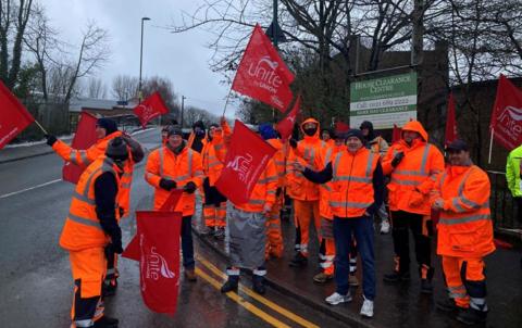 A group of men wearing high-vis orange jackets and trousers stand on a picket line waving red Unite the union flags, next to a road near the canal.