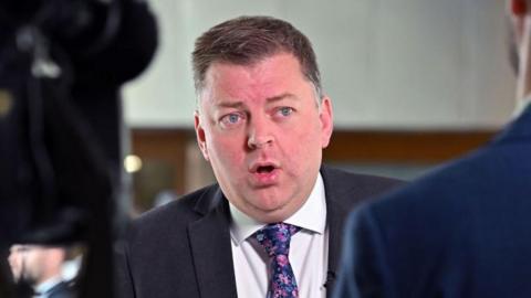 Colin Smyth, a man with spiky brown hair mid speech while wearing a suit, white shirt and blue flowery tie