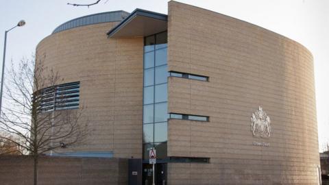 Cambridge Crown Court as seen from outside. It is a cream building that is most likely three storeys high. In the middle it has windows running across each floor in a vertical line. The building looks to be circular in its shape and it has been pictured on a grey day.