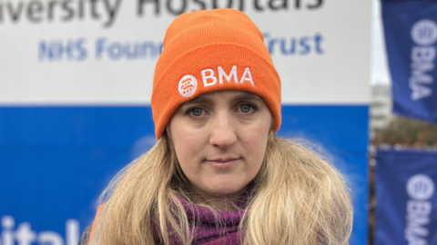 Dr Hanna Copley is unsmiling and looking directly at the camera. She is wearing an orange hat that says BMA on it and is stood in front of a sign. 