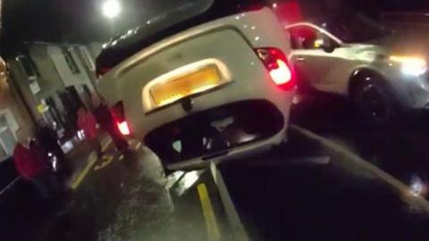 A screenshot taken from a body-worn camera showing a white overturned car in the middle of a road, with four people stood next to it. Their faces are blurred.