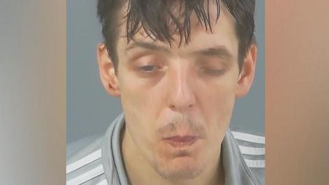 In a custody photo, Ashley Lear has one eye closed and is pursing his lips. He has messy, dark hair and a slight moustache and wears a light grey top with white stripes.