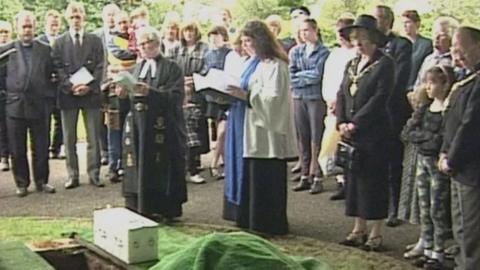 The funeral of baby Callum, showing a small white coffin beside a grave, as mourners gather around clergymen and women reading from books