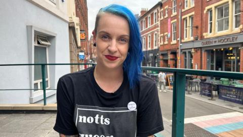 CJ smiles at the camera - they have blue hair and red lipstick. They are wearing a black t-shirt which says the word 'notts' on it.