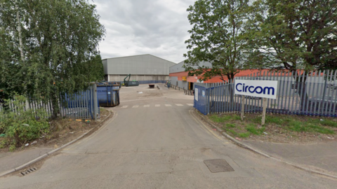 The entrance to a factory, with a white sign that has a blue logo on it which reads Circom. There is blue fencing either side of the entrance and white road markings for a pedestrian crossing along the front of it.