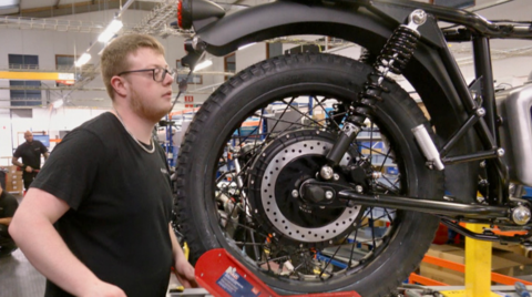 A man with glasses and brown hair is near the back of a motorbike, which is on a raised surface.