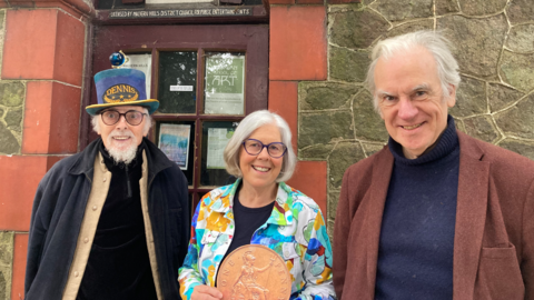 Two men and one woman stood in front of a red brick framed door. One man is wearing a blue top hat with the word "Dennis" on it and the other has white hair and a brown jacket. The woman in the middle is wearing a multi-coloured jacket and is holding an oversized copper coin