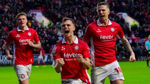 Scott Twine (centre) clenches both his fists and has his mouth open as he shouts in celebration after scoring, with Emil Riis (right) and Cameron Pring (left) behind him