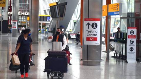 Passengers push their luggage past signage displaying the way to a Covid-19 test centre, in Terminal 5 at Heathrow airport in London,