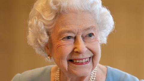 The late Queen. She is smiling and is wearing a duck-egg blue dress, a pearl necklace and pearl earrings.