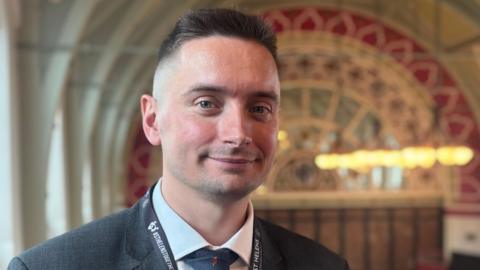 George Woodward, the council leader in St Helens, smiles for the camera. He has short, spikey hair which is shaved on the sides. He is wearing a grey suit and standing inside an ornate building.