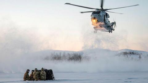 A helicopter is flying over a snowy landscape. Some marines are sitting together on the snow.
