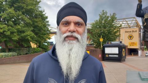 Manny Singh Kang standing in front of the Wolves football stadium. He has a dark head covering, wears a blue top and has a long white beard