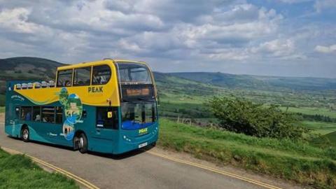 A open top yellow and green bus in the sunshine on a country lane with rolling green hills in the background. 