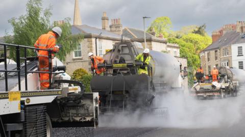 A highways maintenance crew work to maintain a road in a historic-looking town.