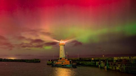 The Northern Lights lighting up the sky in pink and green colours. A white lighthouse and a harbour is at the bottom of the image. 