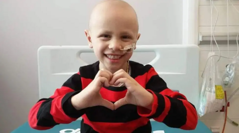 Ben is wearing a black-and-red hooped jumper and forming a heart with his hands. He is in hospital and has a tube inserted into his left nostril. He is smiling.