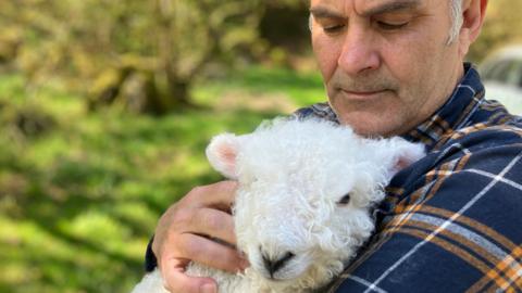 Stephen wearing a check shirt cuddles one of his rare breed lambs on Dartmoor