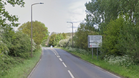 A country road with a sign on the right that reads: HMP Lindholme, HMP Moorland, HMP Hatfield Lakes. There are green bushes, grasses and trees on either side of the road.