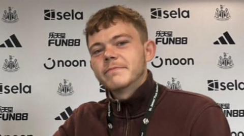 Pardoe Kendrick, wearing a burgundy quarter zip jumper, shaking hands with a man wearing a navy jumper. They are sitting behind a desk at Newcastle United.