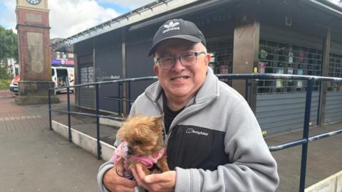 A man holding a dog while smiling. He is wearing a fleece and a black cap while standing outside the corner of a shop close to some railings. 