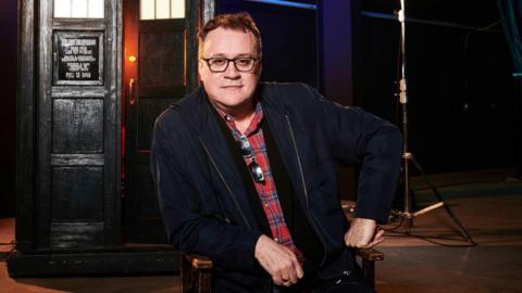 Russell T Davies is wearing a red and blue checked shirt with a black jacket. It's a professional photograph taken on the set of Dr Who. You can see the Tardis in the background. He has dark hair and wears black glasses.