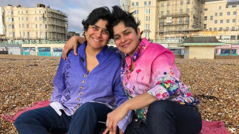 Aisha and Aneesa Chaudhry sit on a beach in Brighton, Aisha wearing a blue top and Aneesa wearing a pink flowery blouse.