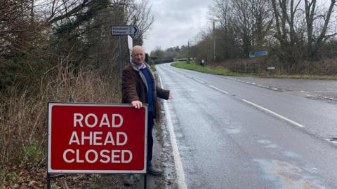A middle-aged man wearing a blue jumper and green jacket stands with his hand on a "road closed" sign in a country road.