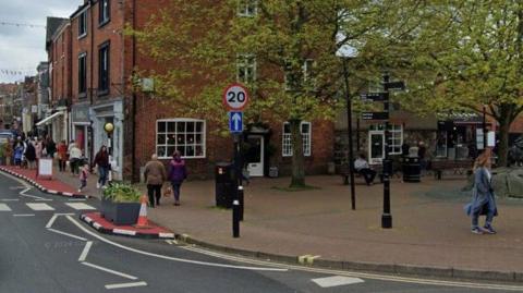 A 20mph speed limit sign on the corner of Church Street, Oswestry. Trees and a red-brick shop building are visible behind, with several shoppers in view. 