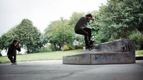 A male skateboarder doing a move on a standalone step. He is being photographed by another skateboarder. Trees and bushes can be seen in the background.