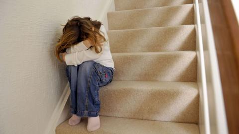 A generic picture of a girl sitting on a carpeted staircase in a home with their head in their hands. The girl is wearing jeans and white sweatshirt, she has long blonde hair but her face is obscured by her posture.