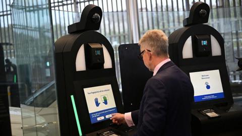 A man with grey hair and wearing a dark suit is entering his personal details into a biometric machine. 