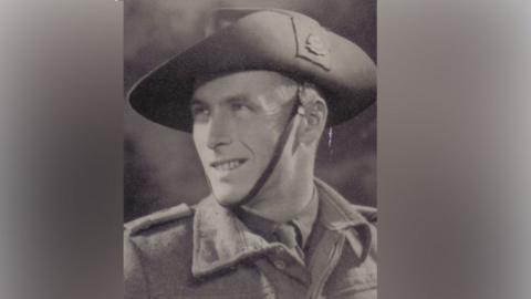 A black and white photo of Jack whose head is turned to the side, looking up slightly with a smile. He wears a wide brimmed hat with a strap under his chin, and a thick army jacket over the top of a shirt and tie.