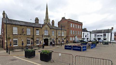 Whittlesey Market Place, showing the George Hotel and a church spire in the backdrop. There are people milling about and outdoor benches to eat on.